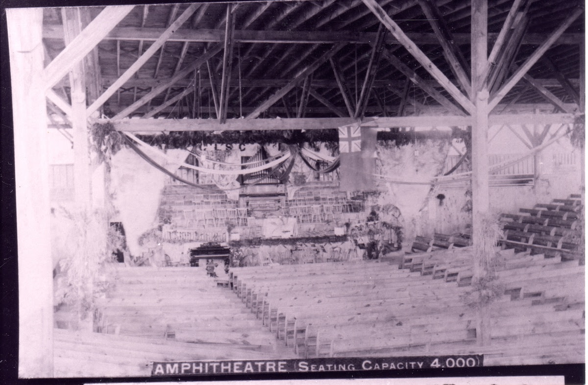 Interior of the open timber Chautauqua amphitheatre: tiered wooden benches descend to a decorated platform with a piano, a Union Jack and a 'C.L.S.C.' banner. A printed caption reads 'Amphitheatre, seating capacity 4,000.'